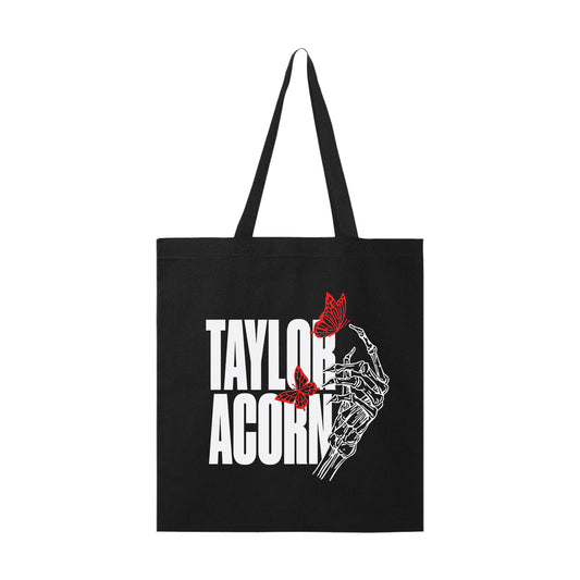 A black tote bag featuring Taylor Acorn in bold white letters and a skeletal hand holding two red butterflies—an eye-catching accessory for any Taylor Acorn fan.