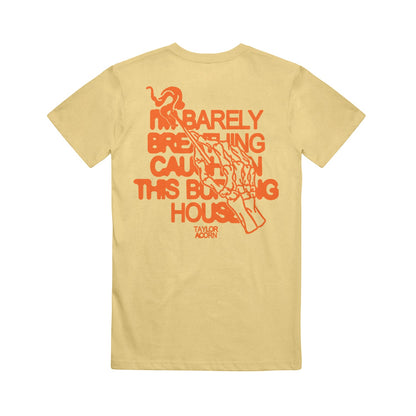 A yellow Taylor Acorn Comfort Colors tee featuring red text on the back: “I’M BARELY BREATHING CAUGHT IN THIS BURNING HOUSE,” with a red skeletal hand holding a match above the brand name.
