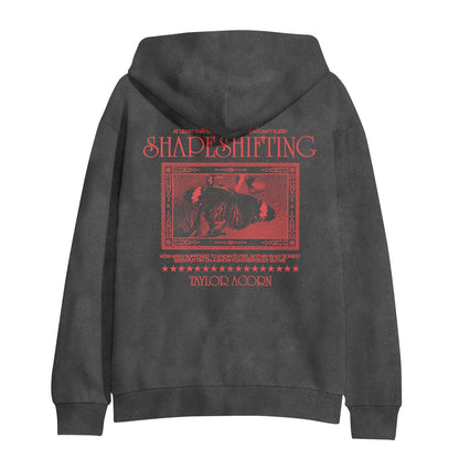 A black Comfort Colors hoodie by Taylor Acorn, featuring bold red graphic text—SHAPESHIFTING and TAYLOR ACORN—above an ornate image, bordered by unique designs and small t-shirt-inspired print on the back.